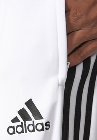 Vita sportbyxor i vitt med svart Adidas-logga och tre vertikala ränder på sidan. Materialet är slätt med en texturerad yta.