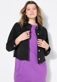 Svart denimjacka med kort längd, silverfärgad kn äppning, framfickor och fransig nederkant, bärs över en lila t-shirt.