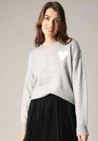 Pull gris en tricot doux, orné d'un motif de cœur blanc sur la poitrine gauche et de poignets côtelés, associé à une jupe plissée noire.