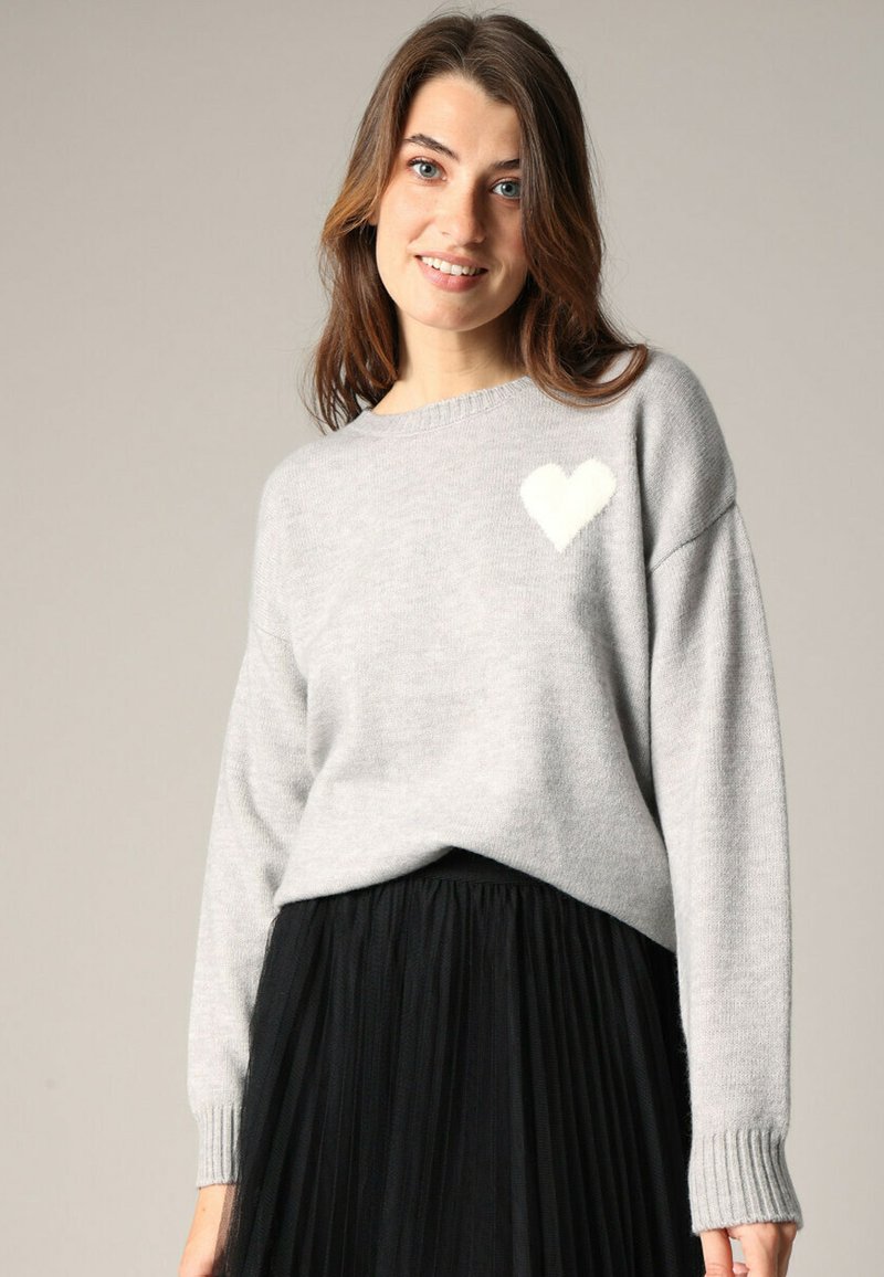 Pull gris en tricot doux, orné d'un motif de cœur blanc sur la poitrine gauche et de poignets côtelés, associé à une jupe plissée noire.