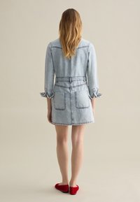 Lichtblauwe denim jurk met lange mouwen, strakke taille en zakken. Heeft opgerolde manchetten en een korte zoom. Gecombineerd met rode ballerina's.
