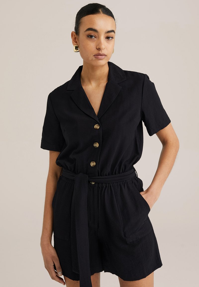 Crna kratka kombinezon s kratkim rukavima, s ovratnikom, prednjim zatvaranjem na pet zlatnih dugmadi, pojasom u struku i dva bočna džepa.