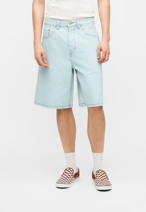 Homme portant un short en jean bleu clair, des chaussettes blanches et des baskets slip-on à carreaux marron et blanc, debout devant un fond blanc.