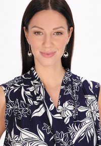 Blusa sin mangas de estampado floral en azul marino con flores blancas. Presenta una tela suave, escote drapeado y una textura lisa. Aretes de aro dorados con perlas.