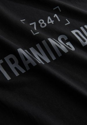 T-shirt en coton noir avec un texte argenté en relief qui indique "ENTRAÎNEMENT PENDANT" et le numéro "7841", avec une texture douce et lisse.