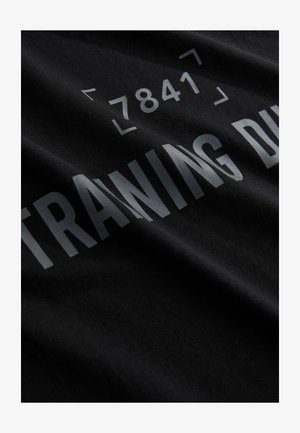 Fekete pamut póló, amelyen kiemelkedő ezüst szöveg olvasható: "EDZÉS KÖZBEN" és a "7841" szám, puha és sima textúrával.