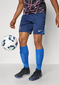 Jūros mėlynos šortai su mėlyna akcentu, mėlynos kojinės ir juodi sportbačiai. Šalia žaidėjo kojų guli balta futbolo kamuolys su juodais ir mėlynais raštais.