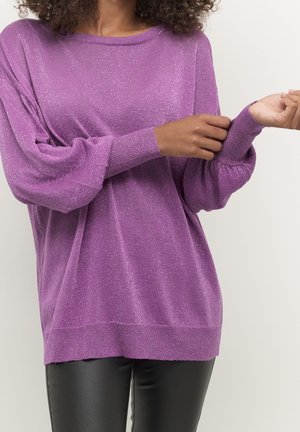 Femme ajustant le poignet d'un pull violet scintillant à manches longues, associé à un pantalon en cuir noir, sur un fond blanc uni.
