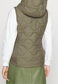 Brązowa pikowana kamizelka z kapturem, ozdobiona falistymi przeszyciami. Noszona na białej koszuli z długim rękawem, w zestawieniu z zieloną skórzaną spódnicą.