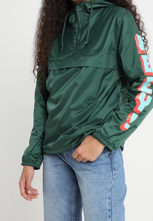 Person iført en mørkegrøn hættetrøje med rød og hvid tekst på ærmet og lyseblå jeans, stående mod en ensfarvet baggrund.