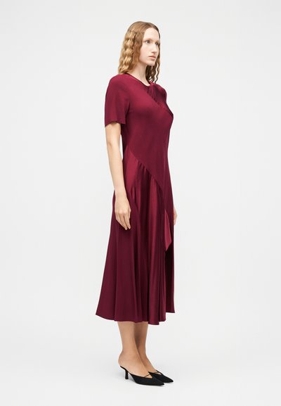 Γυναίκα με σγουρά ξανθά μαλλιά που φοράει midi φόρεμα μπουργκούντι με κοντά μανίκια και ασύμμετρα πάνελ, συνοδευόμενο από μαύρες μυτερές γόβες στιλέτο χαμηλού τακουνιού, στέκεται πλάγια.
