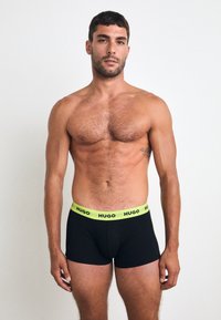 Boxer da uomo neri con cintura in neon verde brillante che presenta il logo "HUGO". Tessuto liscio e vestibilità aderente. Modello in piedi, senza shirt.