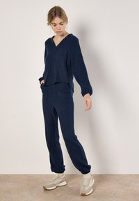 Marineblaues Strick-Loungewear-Set mit einem V-Ausschnitt-Top und elastischen Bündchenhosen, ausgestattet mit zwei seitlichen Taschen und kombiniert mit beigen Sportschuhen.