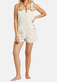 Salopette di denim color crema con spalline regolabili, tasche frontali e dettagli in bottone ottone. Stile shorts con una vestibilità comoda.