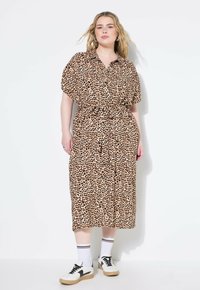 Leopardenmuster-Kleid mit kurzen Ärmeln, Knopfleiste und tailliertem Bund. Der Stoff ist leicht und hat ein fließendes Design. Getragen mit weißen Socken und Turnschuhen.