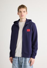 Mørkeblå zip-up hettegenser med en frontlomme og et rødt HUGO-logo-lapp. Brukt over en lys t-skjorte og sammen med lyseblå jeans.