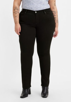 Personne portant un jean noir ajusté, des bottes à talons noires et un haut gris clair à motifs, se tenant avec les bras détendus le long du corps.