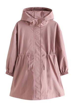 Mladá dievčina stojí, oblečená v mauve kapucňovej bunde, svetložltom topu, svetlomodrých širokých nohaviciach s kvetinovou výšivkou a bielych teniskách.