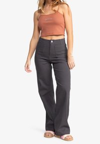Grijze high-waisted broek met rechte pijpen, voorzien van twee voorzakken en een knoopsluiting, gedragen met een cropped roestkleurig tanktop.