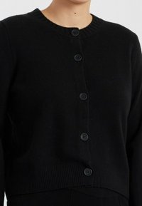 Zwarte gebreide cardigan met een ronde halslijn, geribbelde zoom en zeven zwarte knopen aan de voorkant. Zachte textuur met een getailleerde silhoutte.