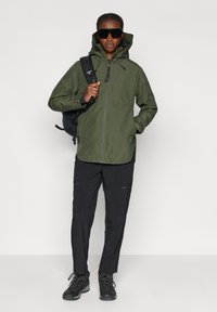 Veste imperméable vert olive avec un col montant, une fermeture éclair à l'avant et une capuche réglable, associée à un pantalon cargo noir et des bottes noires robustes.