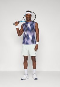 Ärmelloses lila und blau gemustertes Oberteil, helle mintfarbene Shorts, weiße Handgelenkbänder und Schuhe. Hält einen Tennisschläger. Neutraler Hintergrund.