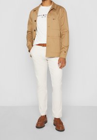 Beige Knopfjacke mit zwei Fronttaschen über einem weißen Logo-T-Shirt, kombiniert mit cremefarbenen Hosen und braunen Slippern.