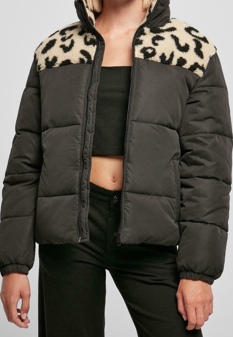 Femme portant une veste matelassée noire avec polaire imprimé léopard aux épaules, ouverte sur un crop top noir et un pantalon noir.