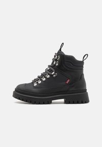 Botas de senderismo negras con suela gruesa, parte superior texturizada, ojales de metal y cordones en contraste blanco y negro. Presentan un asa para facilitar su puesta.