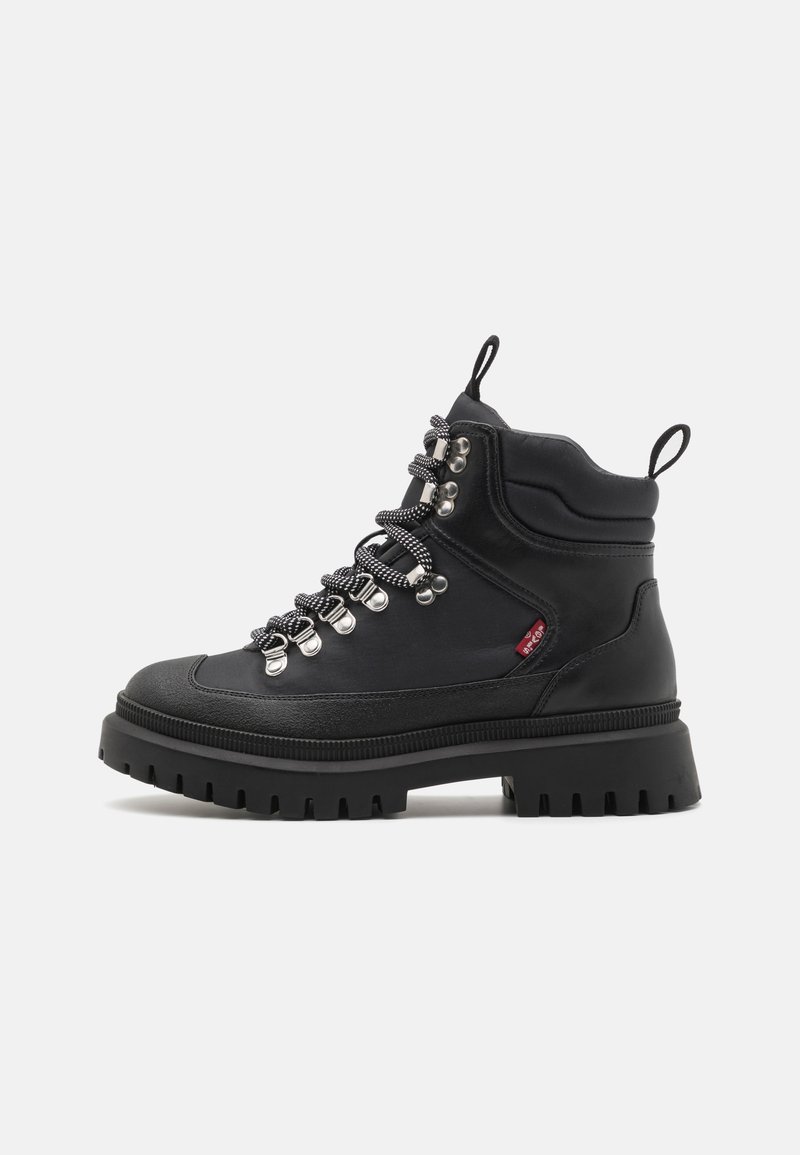 Botas de senderismo negras con suela gruesa, parte superior texturizada, ojales de metal y cordones en contraste blanco y negro. Presentan un asa para facilitar su puesta.
