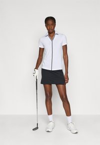 Polo branco de golfe com detalhes pretos, mangas curtas e decote em V; combinado com uma saia preta. A modelo segura um taco de golfe, usando sapatos brancos.