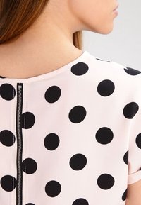 Kortärmad topp i ljusrosa tyg med stora svarta prickar. Har ett blixtlås bak och en rund halsringning.