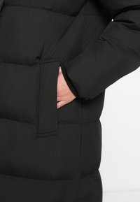 Käsi osaliselt musta, tikitud voodriga jope esitaskus, millel on pikad varrukad.