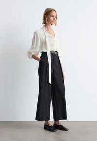 White blouse with a tie at the neck, long sleeves, and a relaxed fit. Black wide-leg trousers with a belt. Black slip-on shoes.