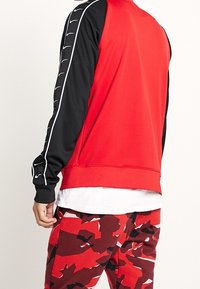 Röd och svart sweatshirt med en mjuk textur, med vita detaljer på ärmarna och en rundad krage. Camo-mönstrade röda byxor.