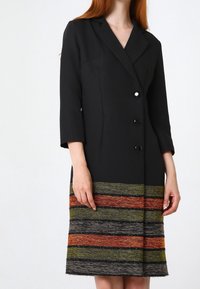 Svart strukturerad klänning med tre framknappar, som har en texturerad fåll i horisontella ränder av grönt, orange och svart.