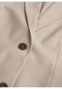 Beżowa marynarka o gładkiej fakturze, z wyciętym klapem, kątowymi szwami i ciemnym guzikiem jako ozdobą.