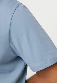 Camisa de manga corta en tela azul clara. Presenta una textura suave y un dobladillo cosido. El primer plano muestra la costura del hombro y la abertura de la manga ligeramente curva.