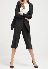 Svart stickad kofta med en draperad silhuett, kombinerad med svarta culottes och spetsiga klackskor. Vit topp syns under.