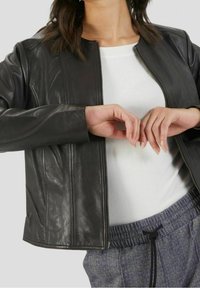 Giacca di pelle nera con zip frontale, cuciture e una texture liscia. Indossata sopra una maglia bianca, abbinata a pantaloni grigi a fantasia.