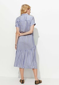 Robe chemise rayée bleu clair avec des manches courtes, ourlet à volants en couches et tissu doux. Portée avec des ballerines noires et beiges à motifs.