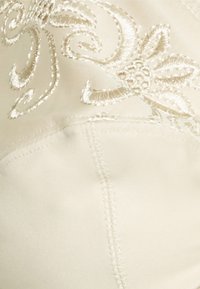 Tela de color crema con un bordado floral blanco intrincado y detalles de costuras texturizadas, que destacan la elegancia y la artesanía.