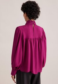 Blusa fucsia con collo alto, dettagli arricciati e maniche lunghe. Presenta una chiusura con bottone sul retro e un tessuto morbido e fluido.