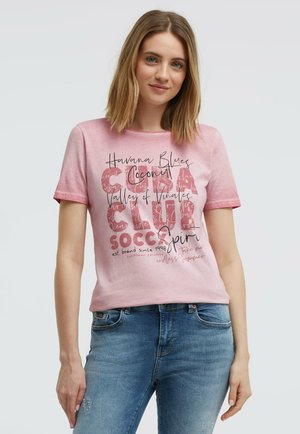 Junge Frau mit schulterlangem Haar, die ein pinkes T-Shirt mit Grafik und blaue Jeans vor einem schlichten hellen Hintergrund trägt.