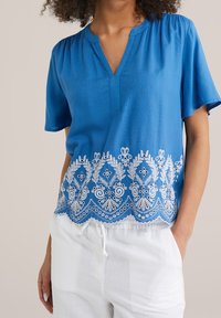 Blå kortärmad blus med V-ringning och dekorativt vitt broderat blommönster vid fållen. Lättviktig tyg med en mjuk textur.