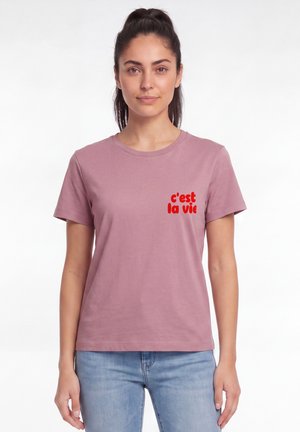 Frau mit dunklem Haar zu einem Pferdeschwanz gebunden, trägt ein mauvefarbenes T-Shirt mit rotem Text "c'est la vie" und hellblaue Jeans, steht vor weißem Hintergrund.