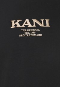 Černé tričko z látky s vyšívaným zlatým textem: "KANI THE ORIGINAL EST. 1989 REG. TRADEMARK," s hladkou strukturou.