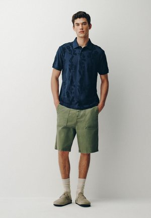 Tânăr care stă cu mâinile în buzunare purtând un tricou polo bleumarin cu imprimeu floral, pantaloni scurți verde măsliniu, șosete bej și pantofi bej pe fundal alb.