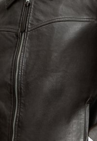 Κοντινό πλάνο σκούρας καφέ δερμάτινου μπουφάν με ορατό ασημένιο φερμουάρ και λεπτομερή ραφή κατά μήκος των ραφών.