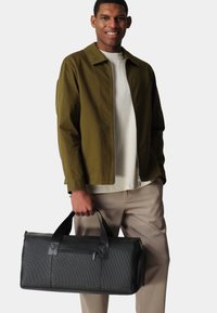 Black textured duffel bag with two sturdy handles, featuring a zippered closure and an external pocket. Worn by a person in casual attire.